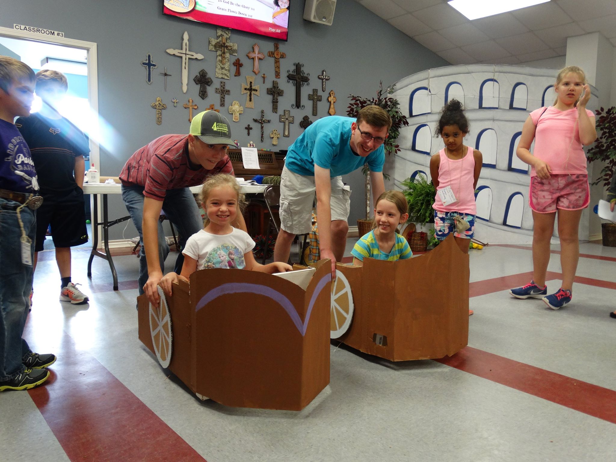 Children gathered inside St. John Lutheran Church Cat Spring preparing to race two decorated cardboard cars, each carrying a young rider.